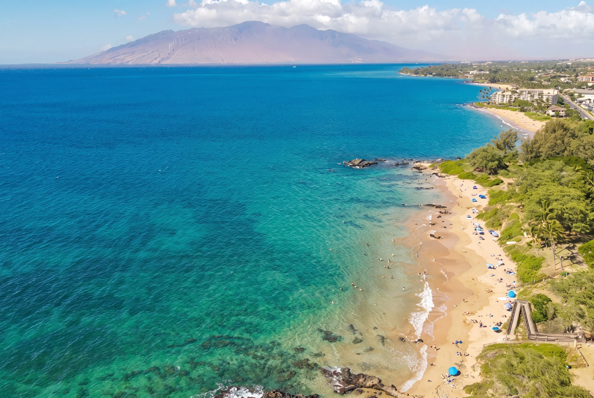Aerial view of the beach and turquoise shallows in South Kihei, with mountains across the bay