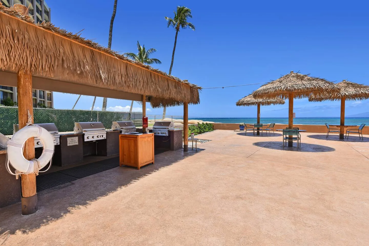 Barbecue grills under a thatched shelter, with tables and umbrellas on the oceanfront deck