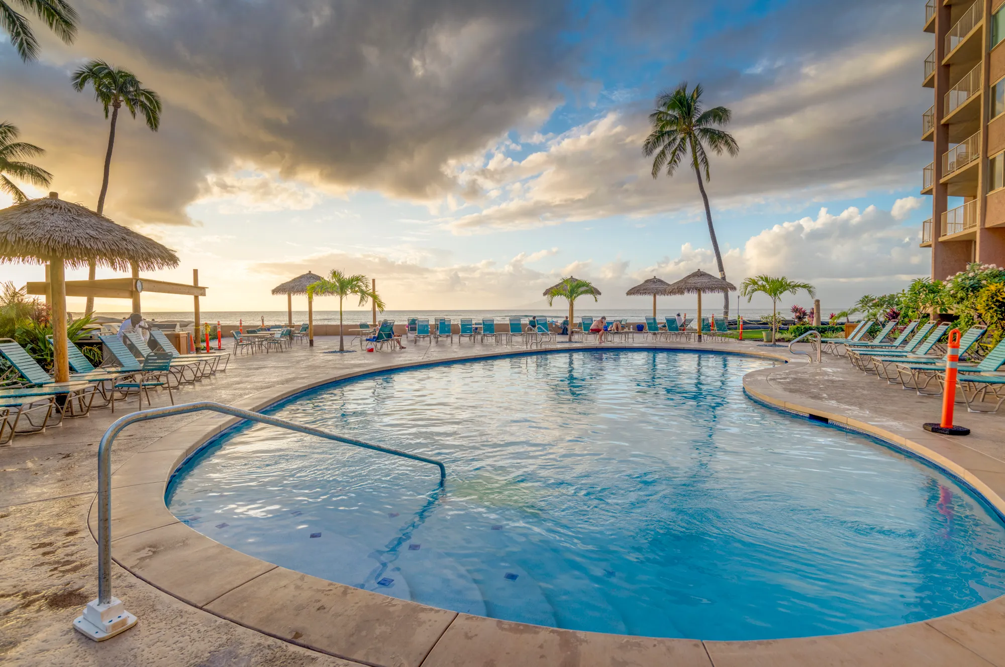 The resort pool looking out to the ocean at sunset, thatched umbrellas along the deck